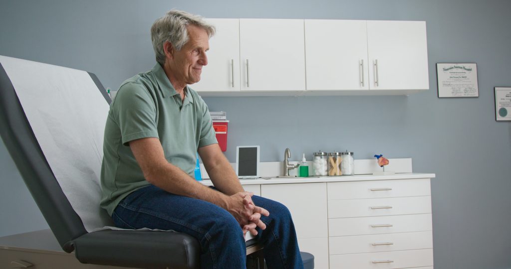 Senior Caucasian male patient waiting patiently for doctor while sitting on exam room table. Older man going to regular appointment for annual check up, low t, clinical research